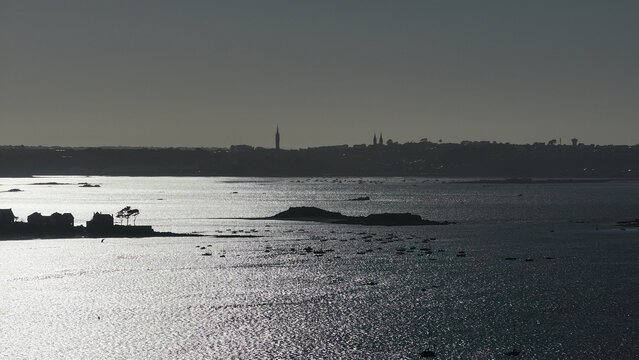 Aerial view of shimmering waters reflect the distant silhouette of the city against the pale sky, creating a serene yet striking contrast, Carantec, Finistre, France.