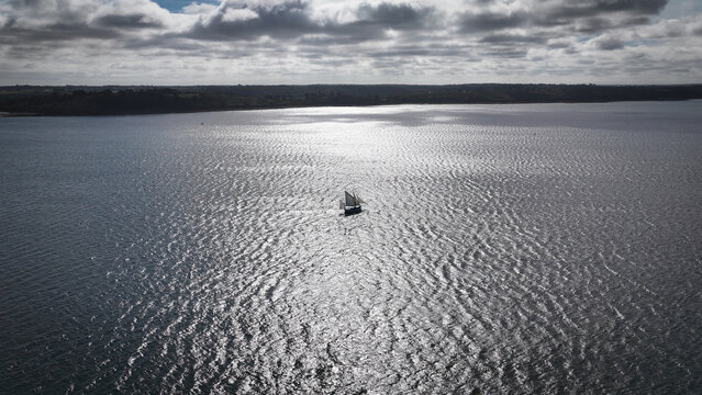 Aerial view of a lone sailboat glides across the shimmering waters, reflecting sunlight amidst the coastal landscape, Carantec, Finistre, France.