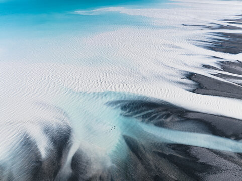 Aerial view of glacial rivers carving through the stark, contrasting landscape with icy blues meeting dark earth, where birds fly over Lake Tekapo, Canterbury Region, New Zealand.