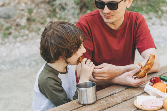 Close-up view of father hugs his school boy son on a family picnic. Child kid and his dad taking a rest and enjoying a picnic while hiking. Boy smiles and bites the bread donut on a picnic.
