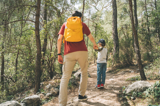 Rear view of tourists school boy and his dad walking a stone footpath in spring forest. Child boy and father wearing casual clothes and yellow backpack while hiking in summer greenwood leaf forest.