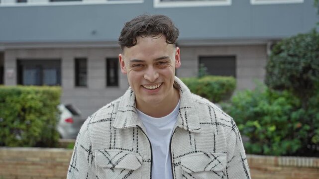 Man in textured white jacket smiling with visible face and slight head tilt in front of a building and green hedges; joy friendship warmth.