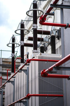 Close-up of power transformers with red fire protection piping and high voltage bushings, showing cooling structures, safety systems, and substation infrastructure outdoors.