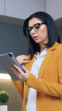 Attractive serious busy 35-aged brunette in eyeglasses dressed in trendy clothes working on tablet pc standing in beautifully decorated cuisine