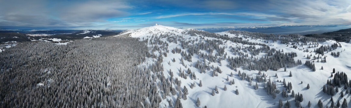 Aerial view of snow-blanketed trees cascade down the mountain slopes towards the town below, a stark white contrast against the dark forest, Mijoux, Auvergne-Rhone-Alpes, France.