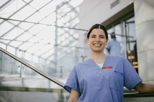 Portrait of smiling female medical expertise wearing scrubs standing near railing in hospital corridor