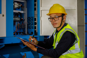 ชายชาวเอเชียวัยผู้ใหญ่ ช่างเทคนิคอุตสาหกรรม สวมหมวกนิรภัยและเสื้อกั๊กนิรภัย ตรวจสอบตู้ควบคุมเครื่องจักรในโรงงาน