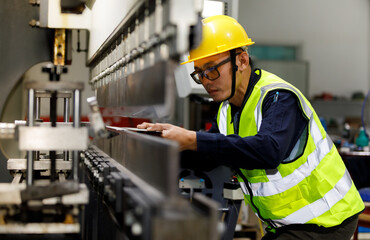 An Asian male industrial engineer, wearing a helmet and safety vest, uses a laptop to monitor the control systems of factory production machinery.