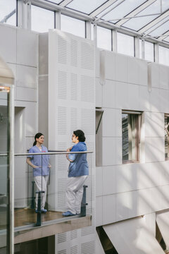Full length of male and female physicians talking with each other while standing in hospital balcony