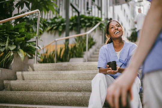 Happy female physician holding coffee mug sitting on stairs while enjoying leisure time with coworker in hospital buildi