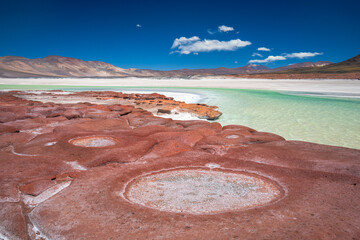 A vibrant panoramic view of a turquoise salt lagoon bordered by red mineral-rich rocks and distant volcanic mountains under a deep blue sky in the Altiplano, Chile. © marcodenaro