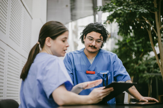 Young male physician listening to female nurse explaining medical documents in hospital
