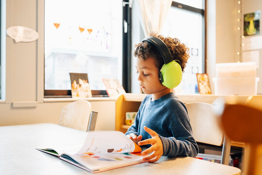 Boy wearing green ear muffs while reading book at table in kindergarten classroom