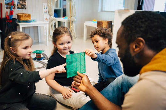 Male teacher holding green translucent shape for group of three curious kindergarten students