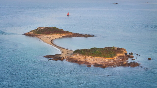 Aerial view of a thin land bridge connecting two rocky, verdant islands amidst the vast, shimmering sea, a solitary sailboat drifting in the distance, Carantec, Brittany, France.