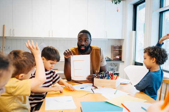 Mature male teacher holding letter E and interacting with students sitting around table in classroom