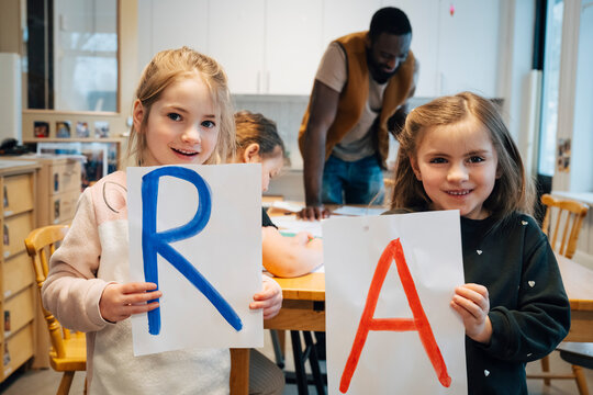 Smiling girls holding papers with letter R and A written on it during alphabet learning game in classroom