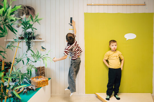 Boy standing against bright yellow backdrop while classmate testing jumping height at preschool