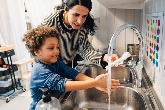 Female teacher assisting boy washing hands in sink after art class at preschool