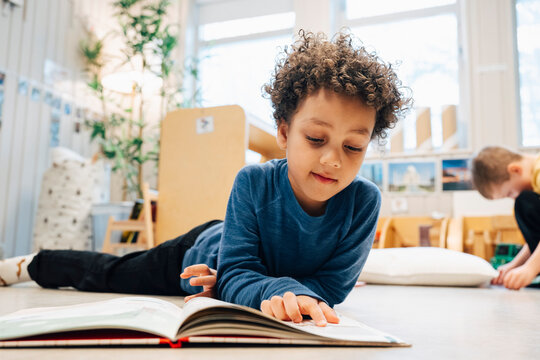 Boy with curly hair reading book while lying on classroom floor