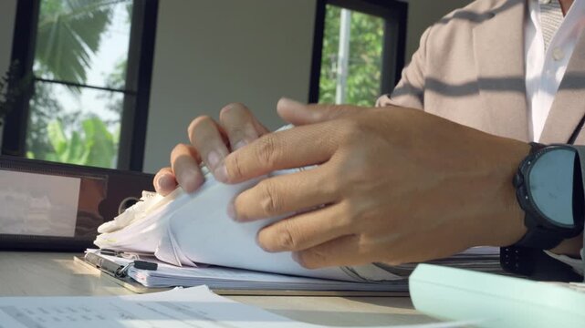 Hands count papers at desk. Natural light streams through window. Businessman wears watch, suit. Documents pile beside black binder. Green foliage visible outside. Fingers move papers, focused