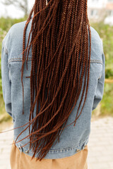 Rear view of a person with long reddish-brown box braids or dreadlocks wearing a blue denim jacket outdoors, close up for hairstyle, ethnic beauty, fashion, and hair care concept. vertical