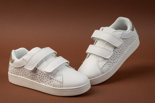 Stylish white kids' sneakers with velcro straps on brown background. Children's fashion footwear for active play and back to school concept.
