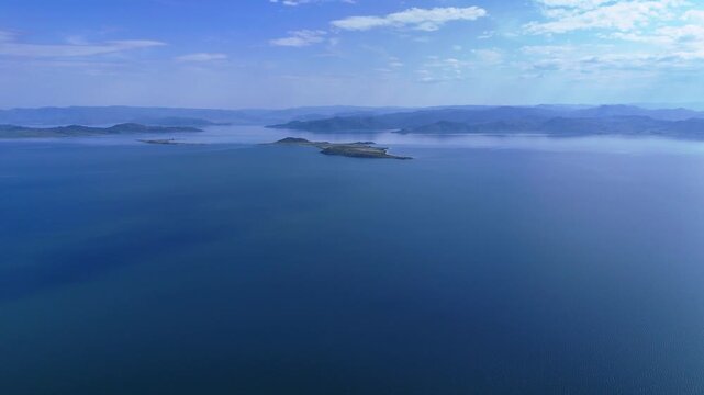 Majestic drone view of the Bukhtarma Reservoir featuring a central island against hilly shores shrouded in a blue haze. Bright, dramatic sunlight pierces through thick clouds, creating a unique