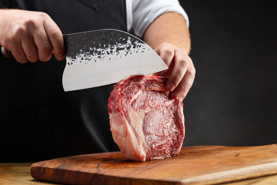 Professional butcher, chef cuts raw beef steak with sharp big knife on a wooden board and wooden table.