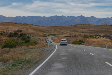 Fototapeta premium Winding Road Through Arid Hills with Distant Mountains Under Cloudy Sky