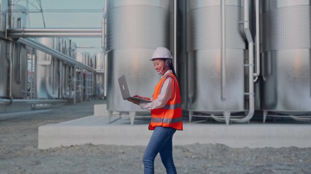 Side View of Asian Female Engineer Use Computer While Walking at Industrial Chemical Plant with Row of Stainless Steel Storage Silos