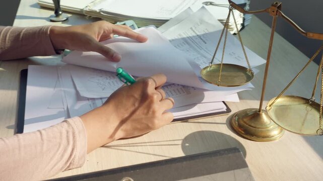 Lawyer reviews documents with a golden scale beside them. Hands hold papers and a green pen over legal paperwork. Desk holds stacks of files and a classic justice symbol