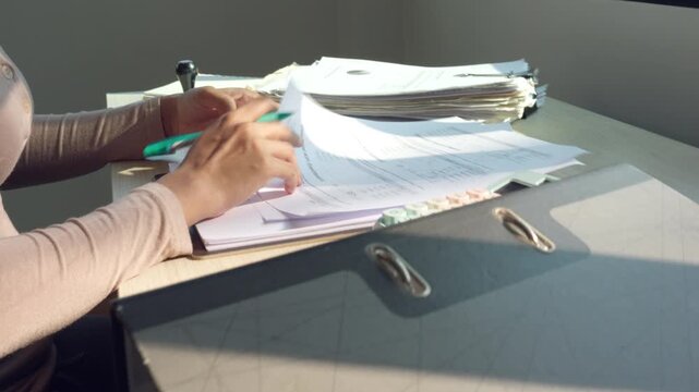 Hands review papers at a cluttered desk. Calculator sits nearby, unused. Sunlight glints across the surface. Hands hold a green pen, poised. Documents stack in soft, natural light
