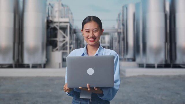 Asian Female With Her Laptop at Chemical Plant with Row of Stainless Steel Storage Silos, She Is Looking At The Camera With A Smile