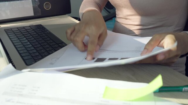 Hand reviews documents while typing on a laptop. Desk holds papers, sticky notes, and a gold scale. Focus on hands and desk items, no animals or plants