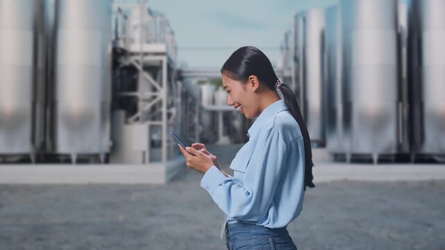 Side View Of Asian Female With Her Smartphone at Chemical Plant with Row of Stainless Steel Storage Silos, Checking On Her Smartphone With Meditation