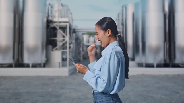 Side View Of Asian Female Professional Worker With Her Smartphone at Chemical Plant with Row of Stainless Steel Storage Silos, She Raises Her Fist Up With Screaming Goal After Check On The Smartphone