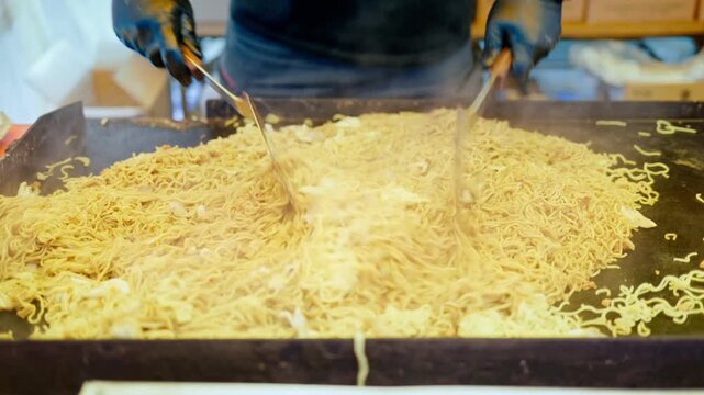 Close-up of noodles being stir-fried on a large griddle by a chef using spatulas.