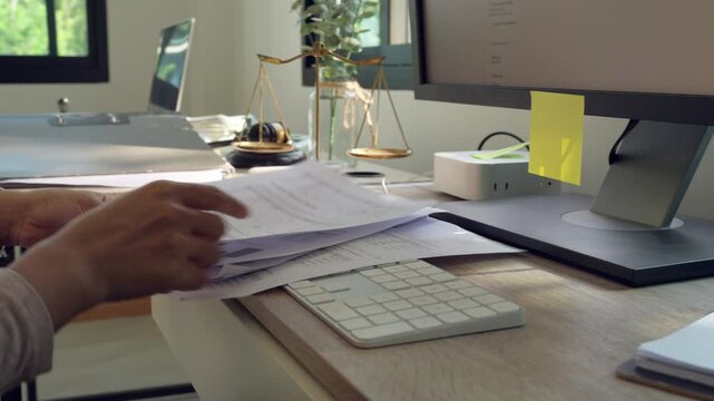 Hands sort papers at a tidy desk. Keyboard and monitor sit beside documents. Natural light streams through a window. Small plant adds green to the workspace. Desk holds a scale and yellow sticky note