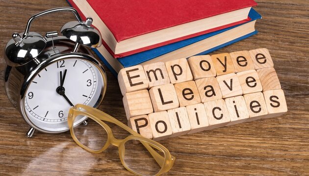 Wooden cubes spelling &ldquo;Employee Leave Policies" near with clock, glasses and books on desk