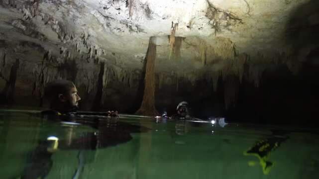 Group of scuba divers emerging in a dark underground cave with stalactites illuminated by flashlights