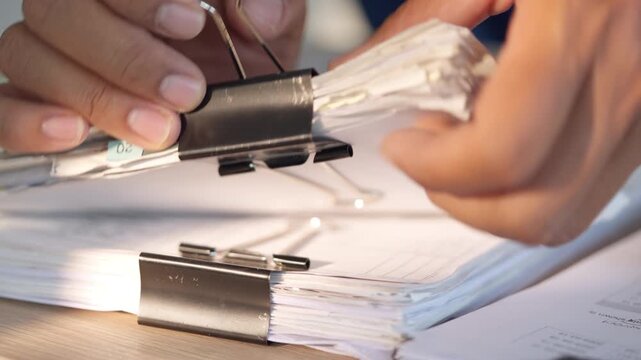 Hands hold papers clipped with metal fasteners. Fingers adjust documents stacked on a flat surface. Sunlight glows softly through the workspace. Office tasks appear precise and focused