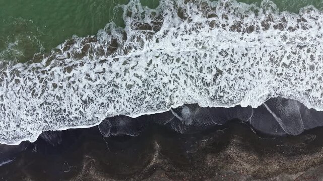 Waves roll onto a dark sand beach. The white foam of the water contrasts with the dark sand. The sun shines brightly over the scene, creating clear visibility of waves and shore.