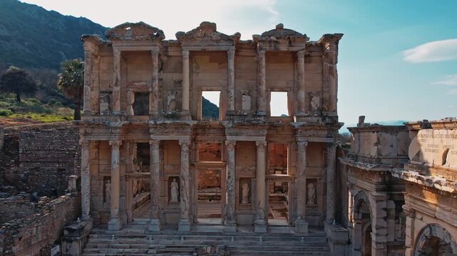 Wide cinematic view of the Library of Celsus in the ancient city of Ephesus, Izmir, Turkey. Iconic Roman ruins, historic architecture and travelers under clear daylight sky and palm trees.