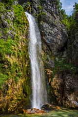 Obraz premium A majestic Grunas Waterfall dramatically cascades vertically over a high, textured rock cliff adorned with lush green moss and foliage in Theth, Albania.