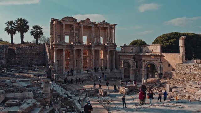 Wide cinematic view of the Library of Celsus in the ancient city of Ephesus, Izmir, Turkey. Iconic Roman ruins, historic architecture and travelers under clear daylight sky and palm trees.