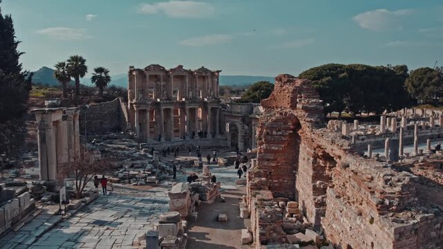 Wide cinematic view of the Library of Celsus in the ancient city of Ephesus, Izmir, Turkey. Iconic Roman ruins, historic architecture and travelers under clear daylight sky and palm trees.