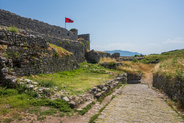 A historic cobblestone walkway winds between the rugged, ancient stone walls of Rozafa Castle, highlighting its formidable defenses under a bright summer sky in Shkoder, Albania. © Danica Chang