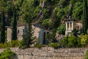 Obraz premium An ancient stone church with arched windows and an adjacent bell tower, topped with a cross, stands amidst lush green trees and rocky terrain on a bright day in Berat, Albania.