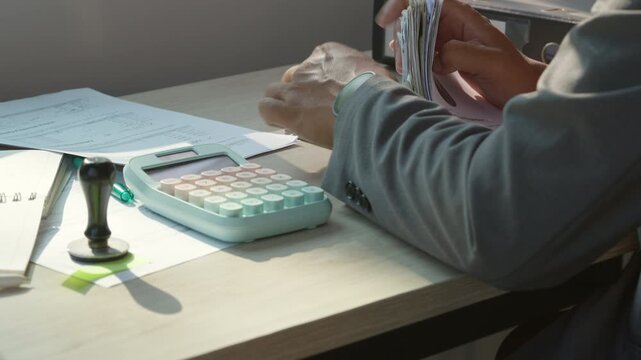 Hands count money at desk with calculator nearby. Office clutter includes papers and stamps. Sunlight falls on workspace, highlighting details. Person wears grey suit jacket while working